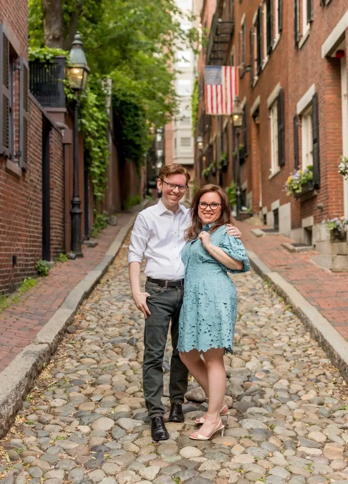 A smiling man and woman standing together on a cobblestone street in an alleyway, surrounded by brick buildings and greenery, with an American flag hanging from a building.