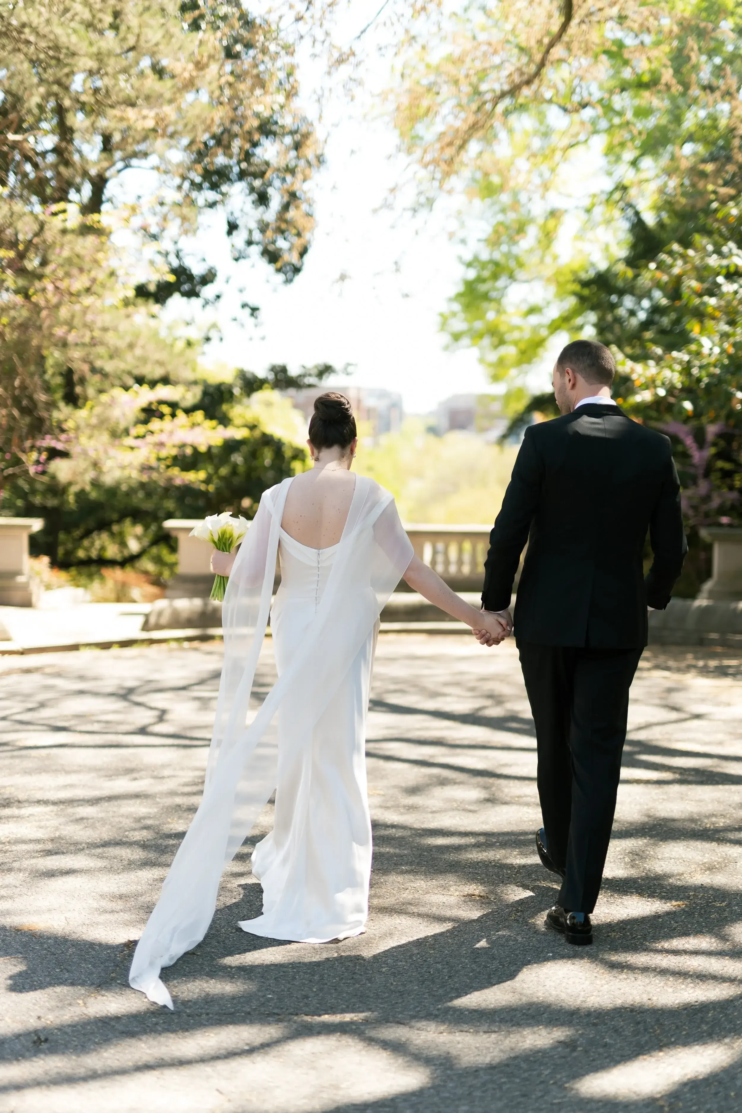 A bride and groom holding hands walking outdoors on a sunny day with trees in the background.