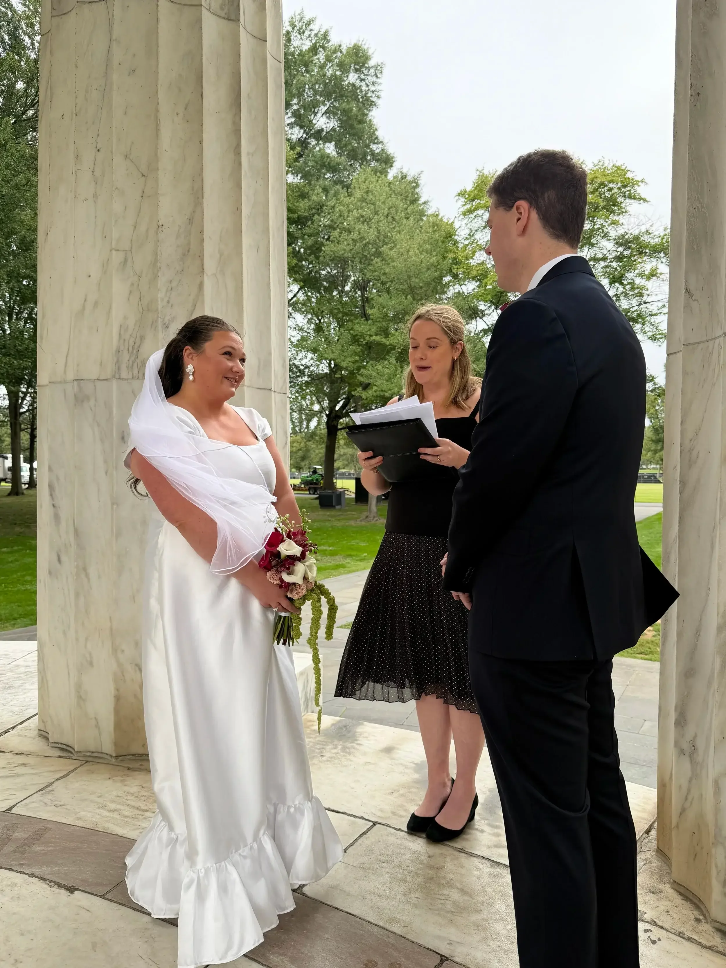 A bride and groom standing at an outdoor wedding ceremony, with an officiant holding a binder, in front of marble columns and green trees.