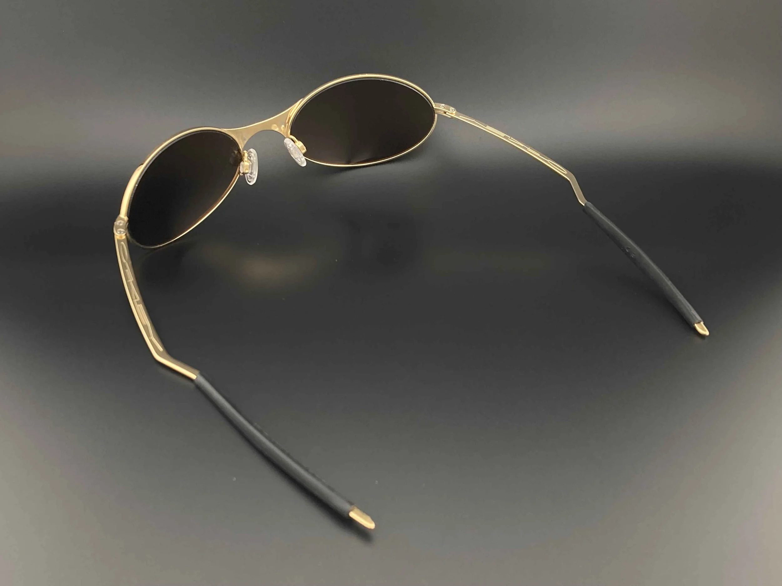 Pair of sunglasses with gold frames, dark oval lenses, and black temples on a dark surface.
