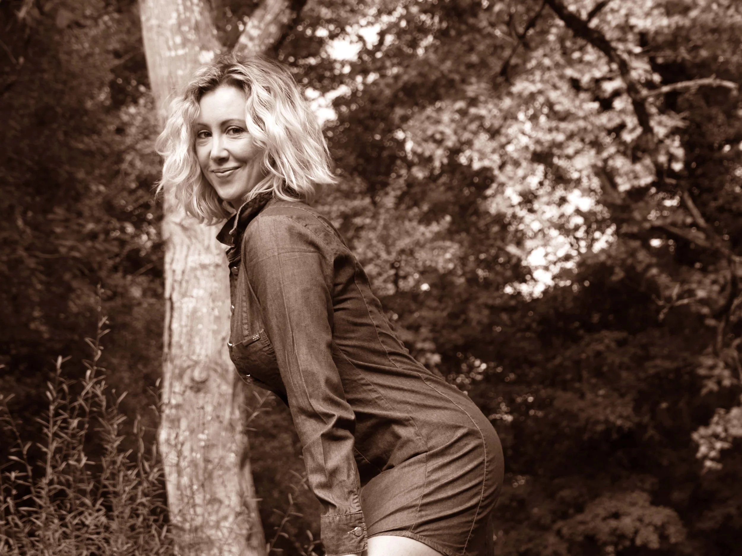 Femme blonde souriante, portant une chemise en jean, se tenant dans un cadre naturel avec des arbres en arrière-plan.