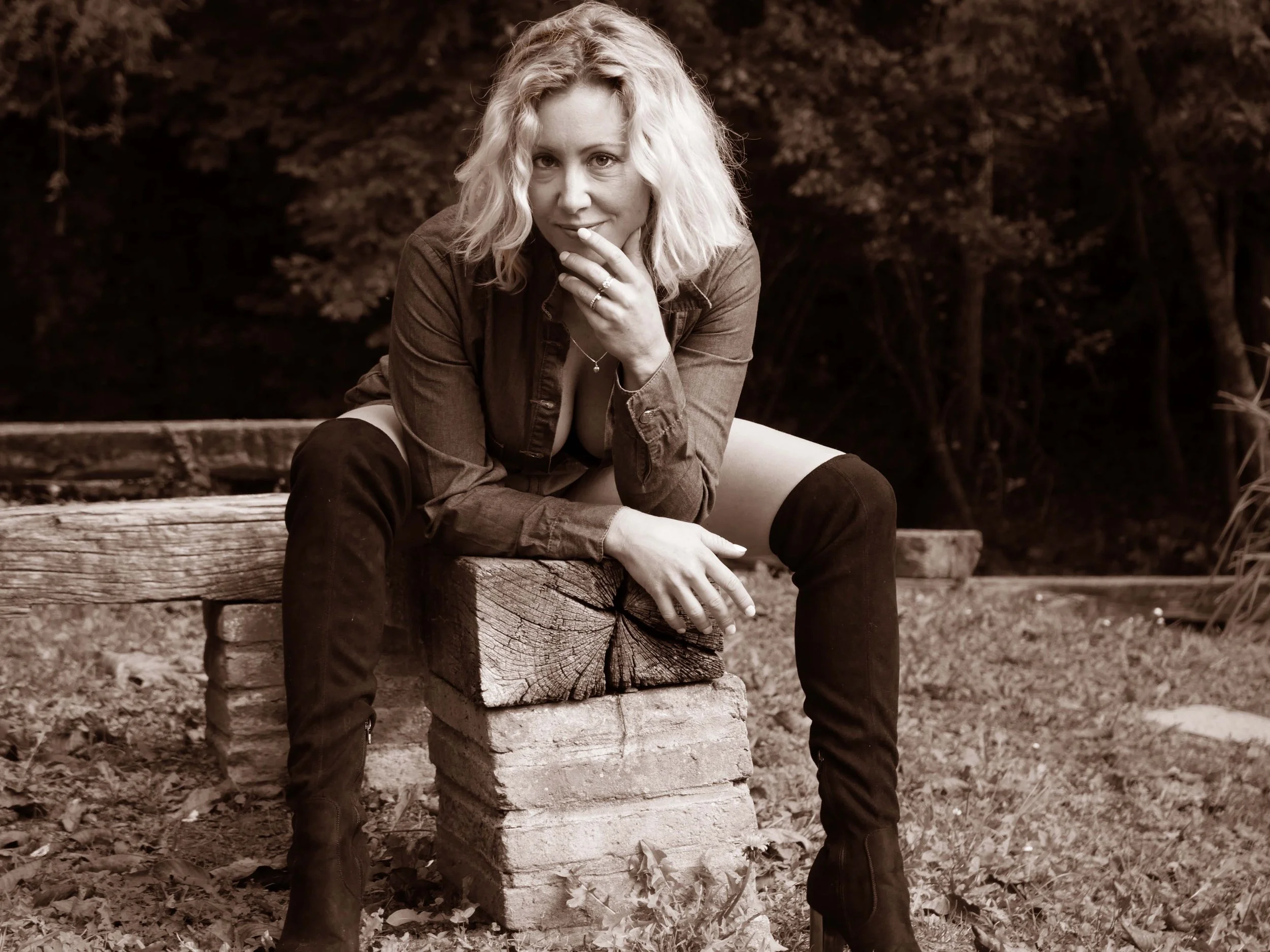 Une femme aux cheveux blonds assise sur une bûche en bois, dans un environnement extérieur, portant des bottes hautes et une chemise ouverte. Image en noir et blanc.