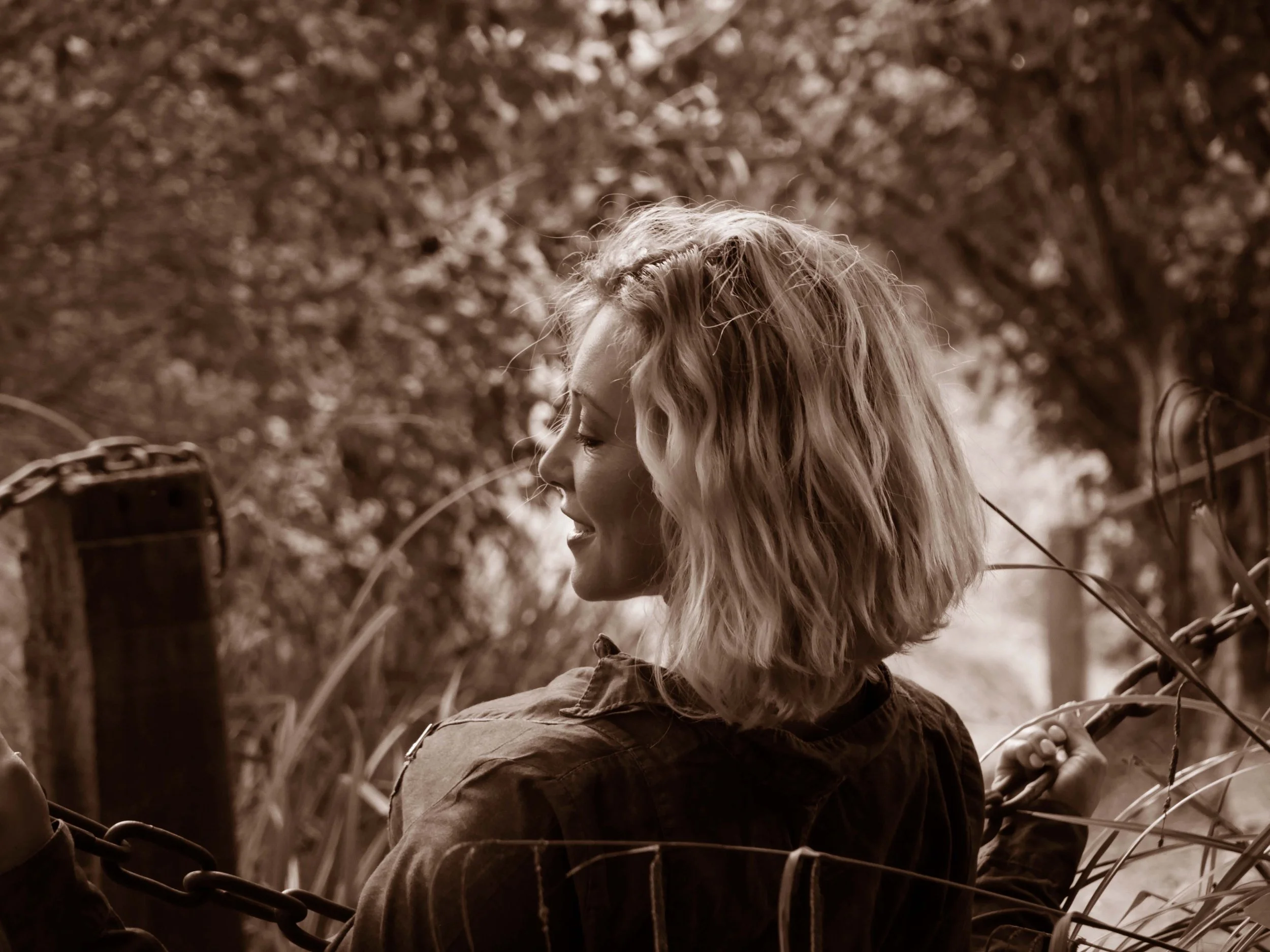 Portrait en sépia d'une femme blonde souriant, tenant une chaîne dans un environnement naturel avec des arbres.