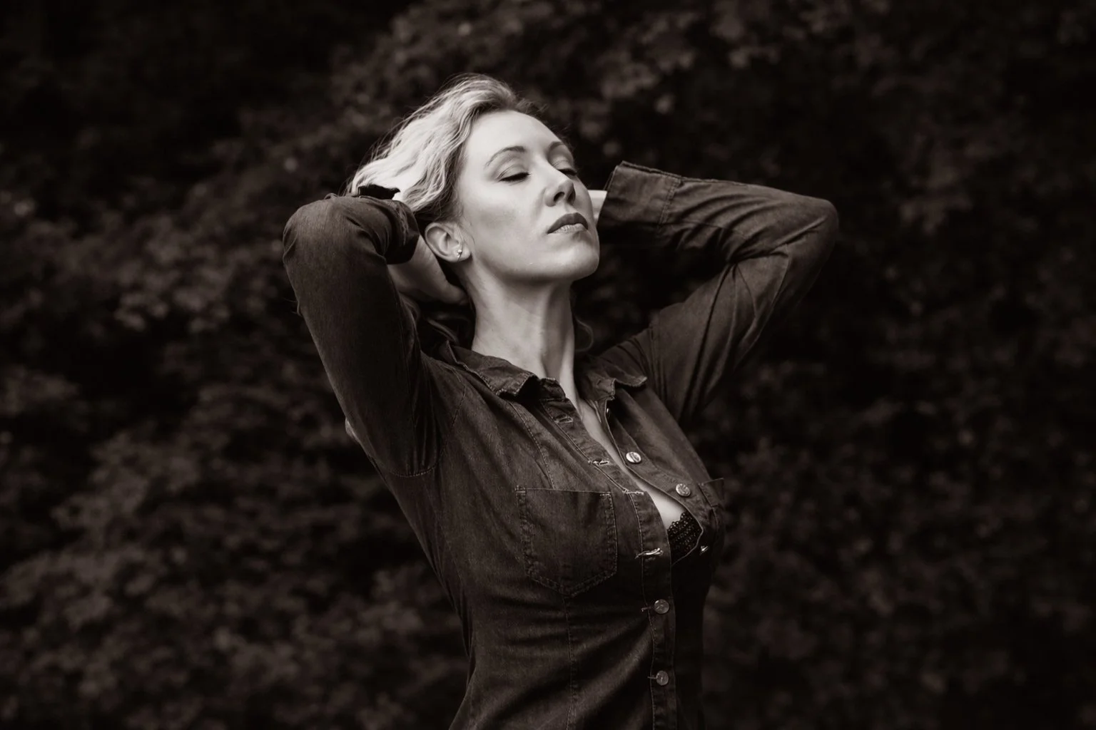 Une femme avec les yeux fermés, les bras derrière la tête, debout dans un environnement naturel, en noir et blanc.