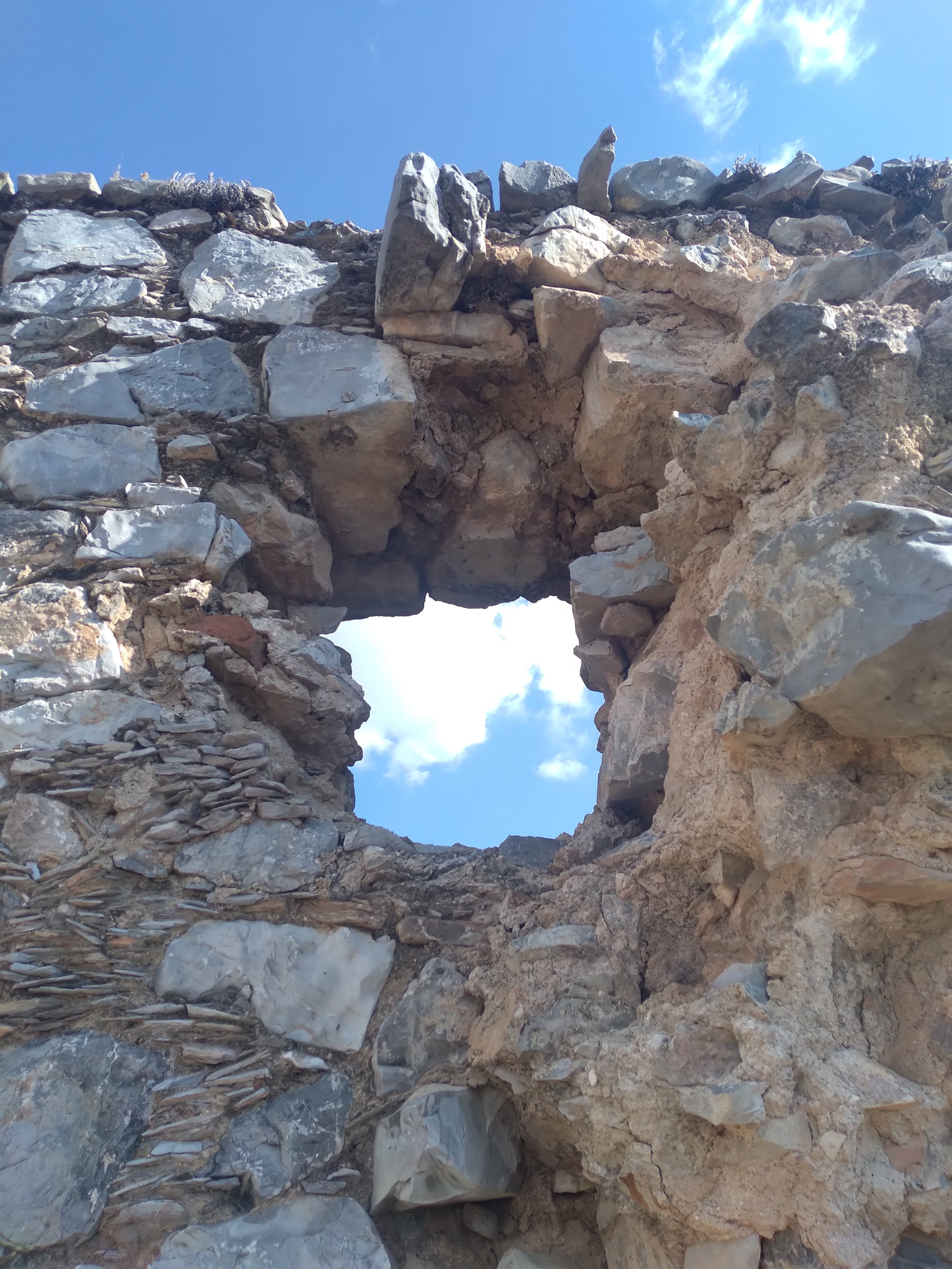 A close-up of a weathered stone arch in a rocky wall with a clear blue sky with some clouds visible through the opening.