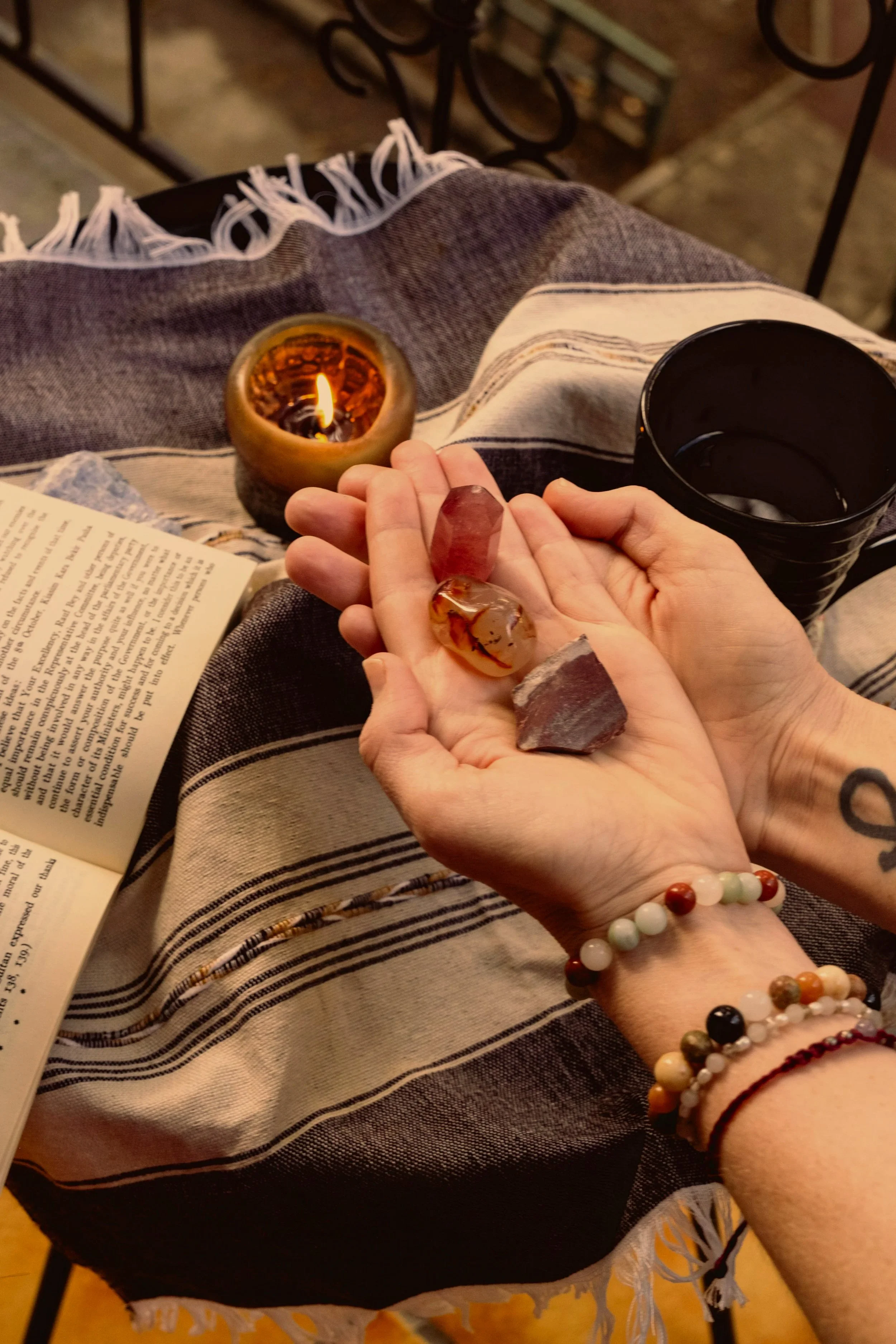 A person holding a handful of colorful gemstones over a striped cloth. On the table, there is a lit candle, an open book, and a black cup. The setting appears cozy and relaxing.