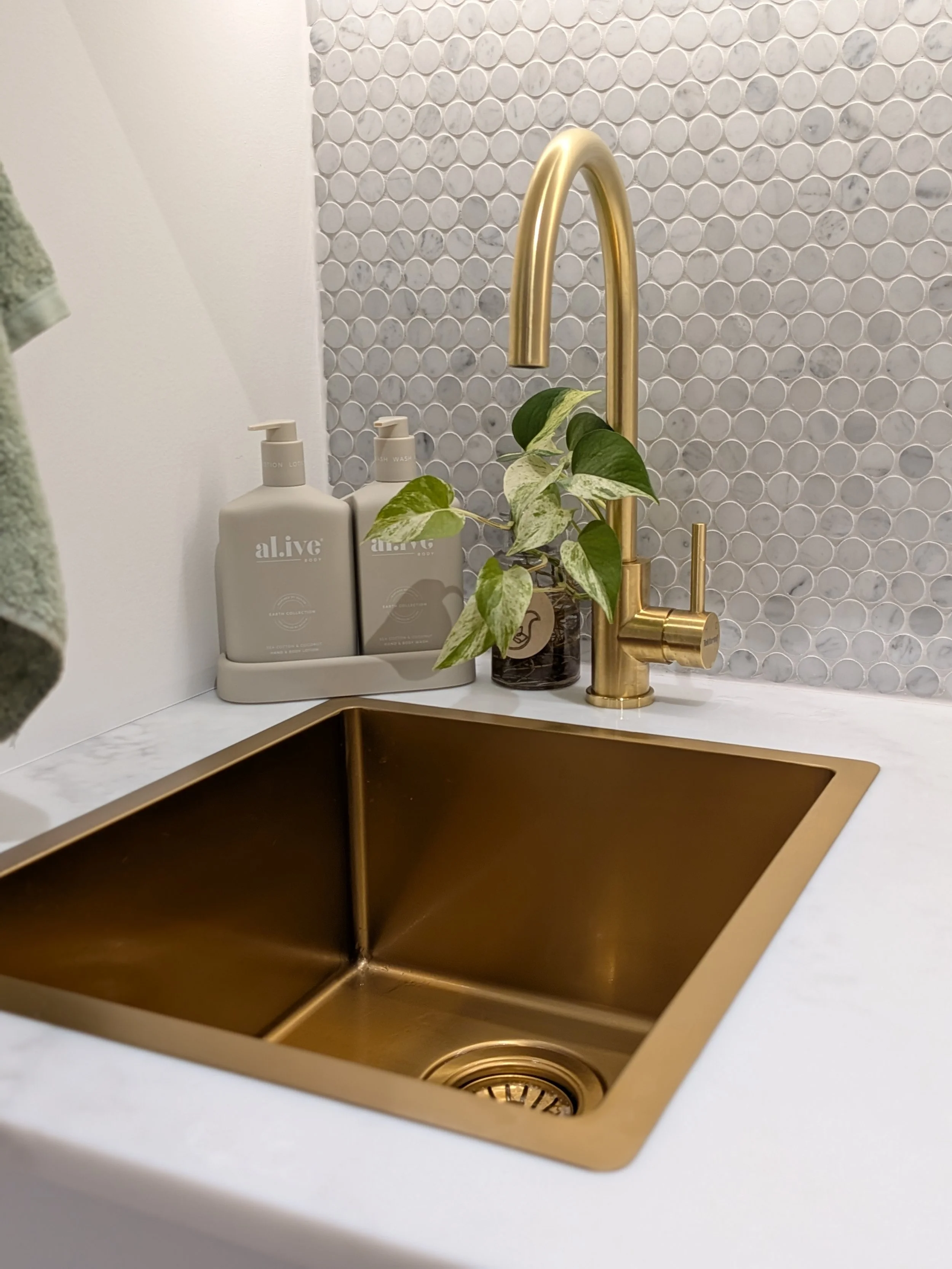 A kitchen sink with a gold-colored faucet, a potted plant, and two bottles of lotion or soap on a white countertop. The background features a white tiled wall with a circular pattern.