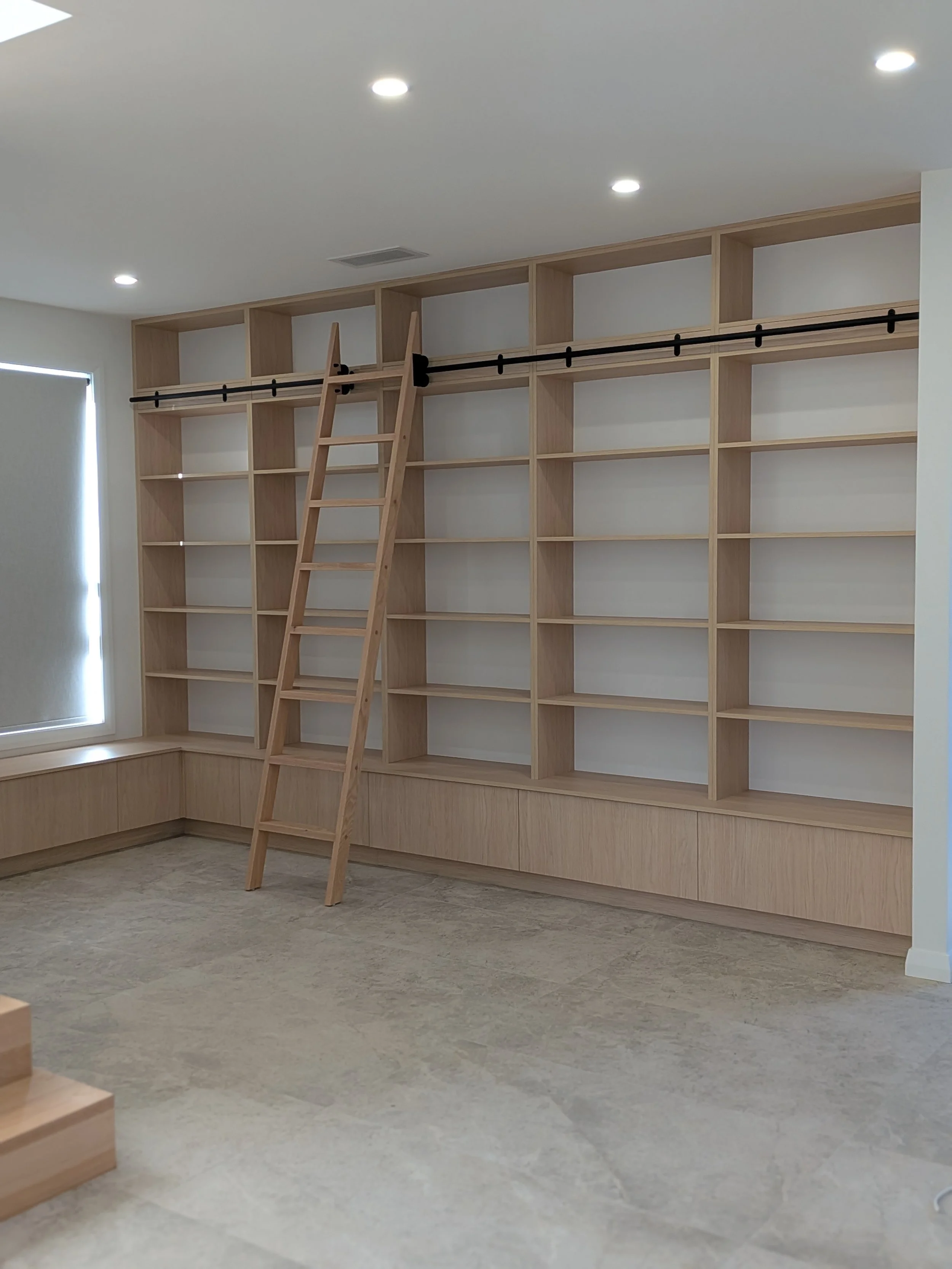 Empty built-in wooden bookshelf with a ladder and black sliding track in a room with white walls, ceiling lights, and a window with closed blinds.