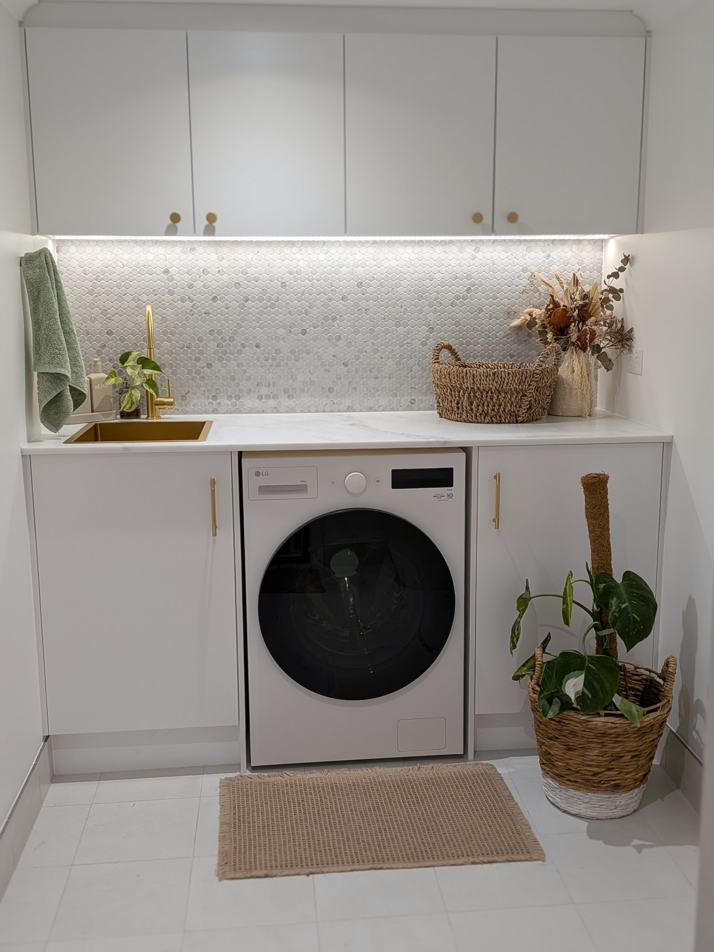 A compact laundry area with a front-loading LG washing machine underneath white cabinets. The countertop has a gold sink with a gold faucet, and a green towel hangs on the wall. A woven basket with dried flowers and a potted plant in a wicker basket sit on the counter. A beige rug is on the white tiled floor.