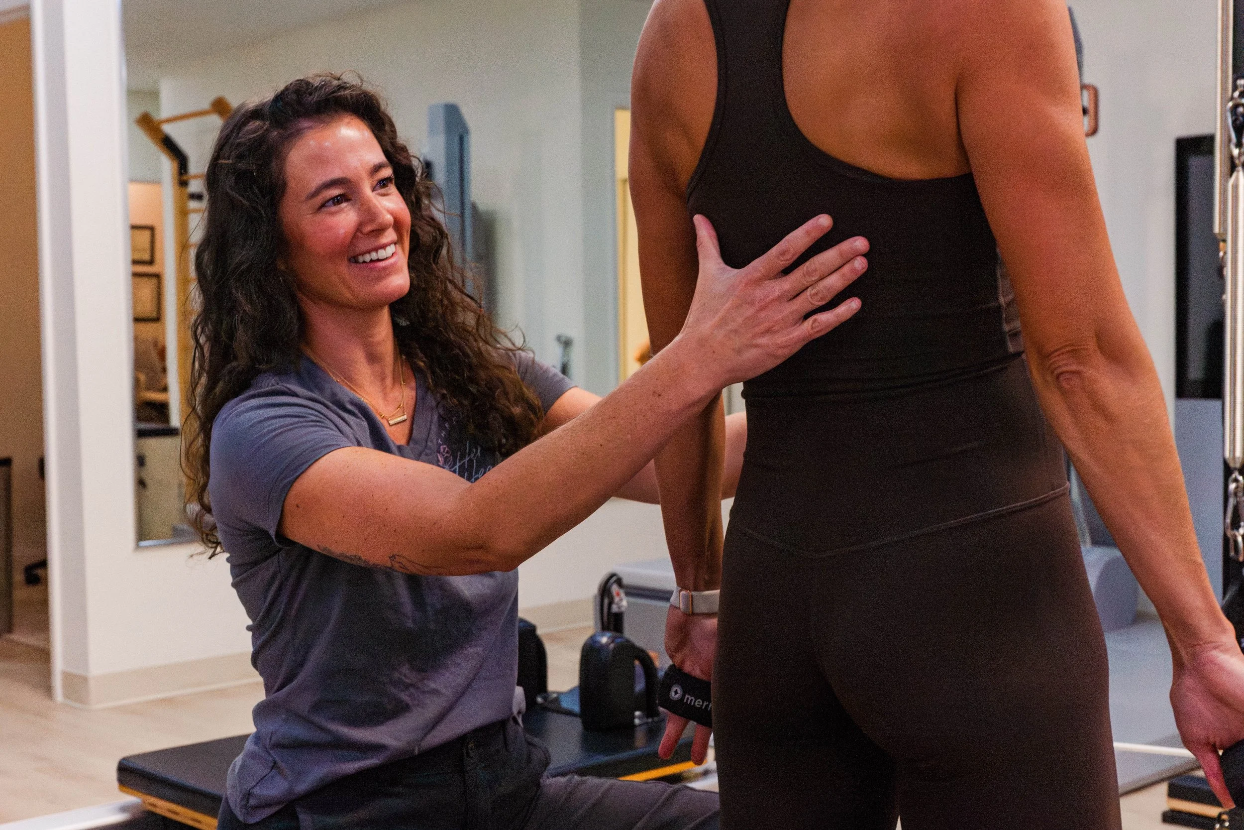 physical therapist giving cues to a patient during a pilates reformer session. patient is in tall kneeling pulling on straps for posture