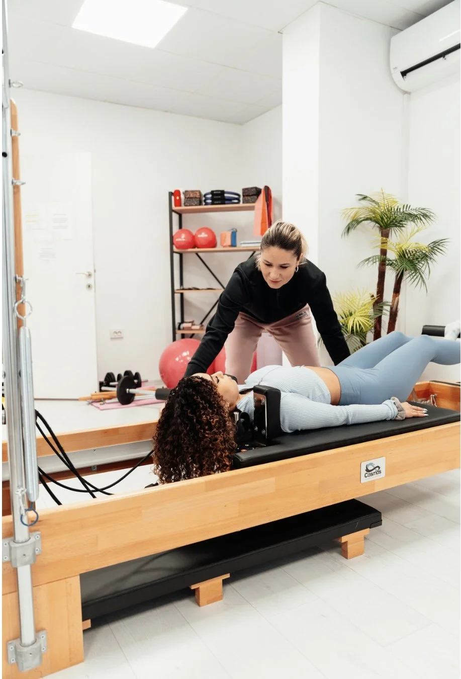 A woman lying on a reformer with a trainer helping her through a personalized 1:! Pilates session.