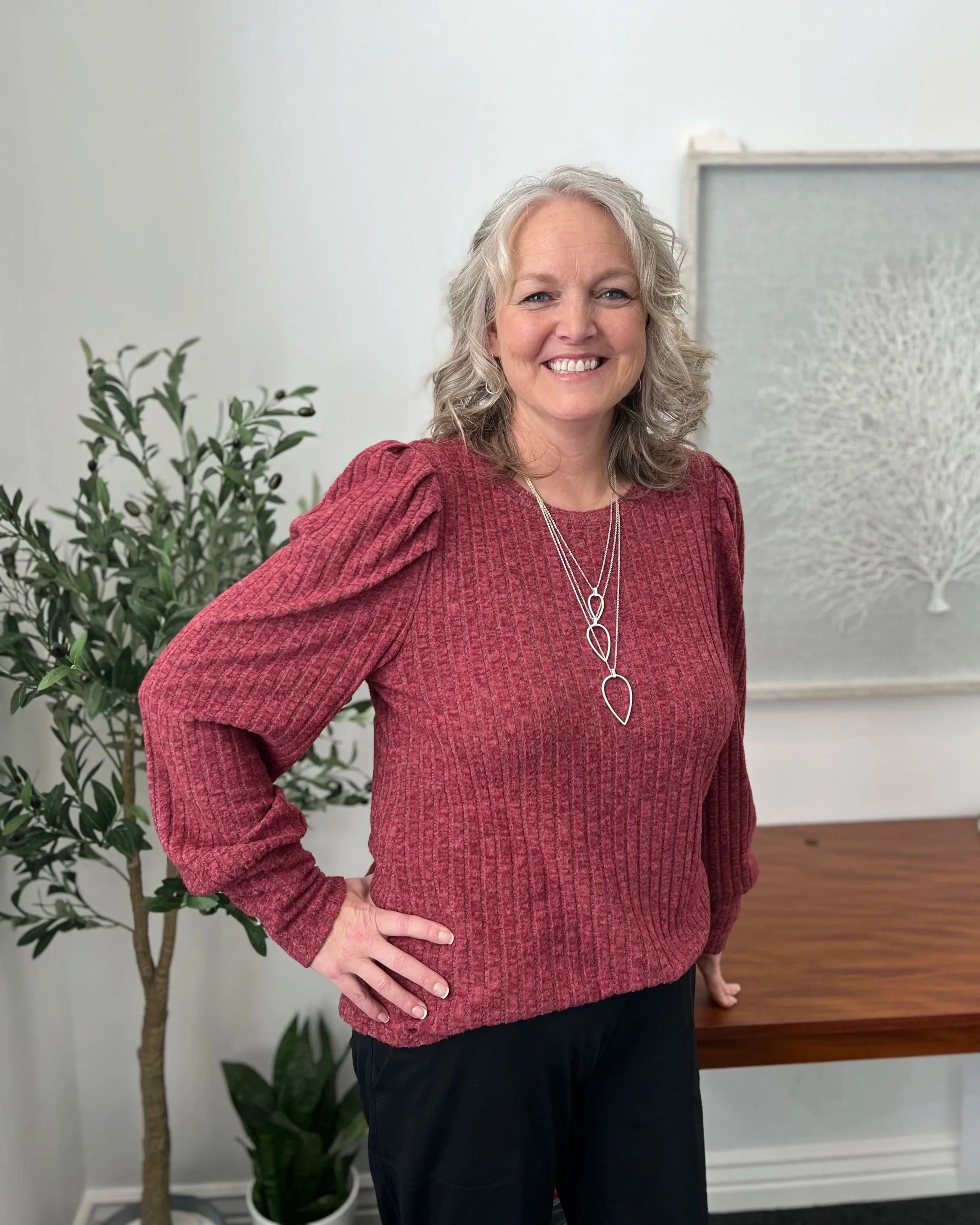 A woman smiling, wearing a red textured sweater with puffed shoulders and a silver necklace, standing in front of a white wall with a framed artwork and a potted plant with green leaves.