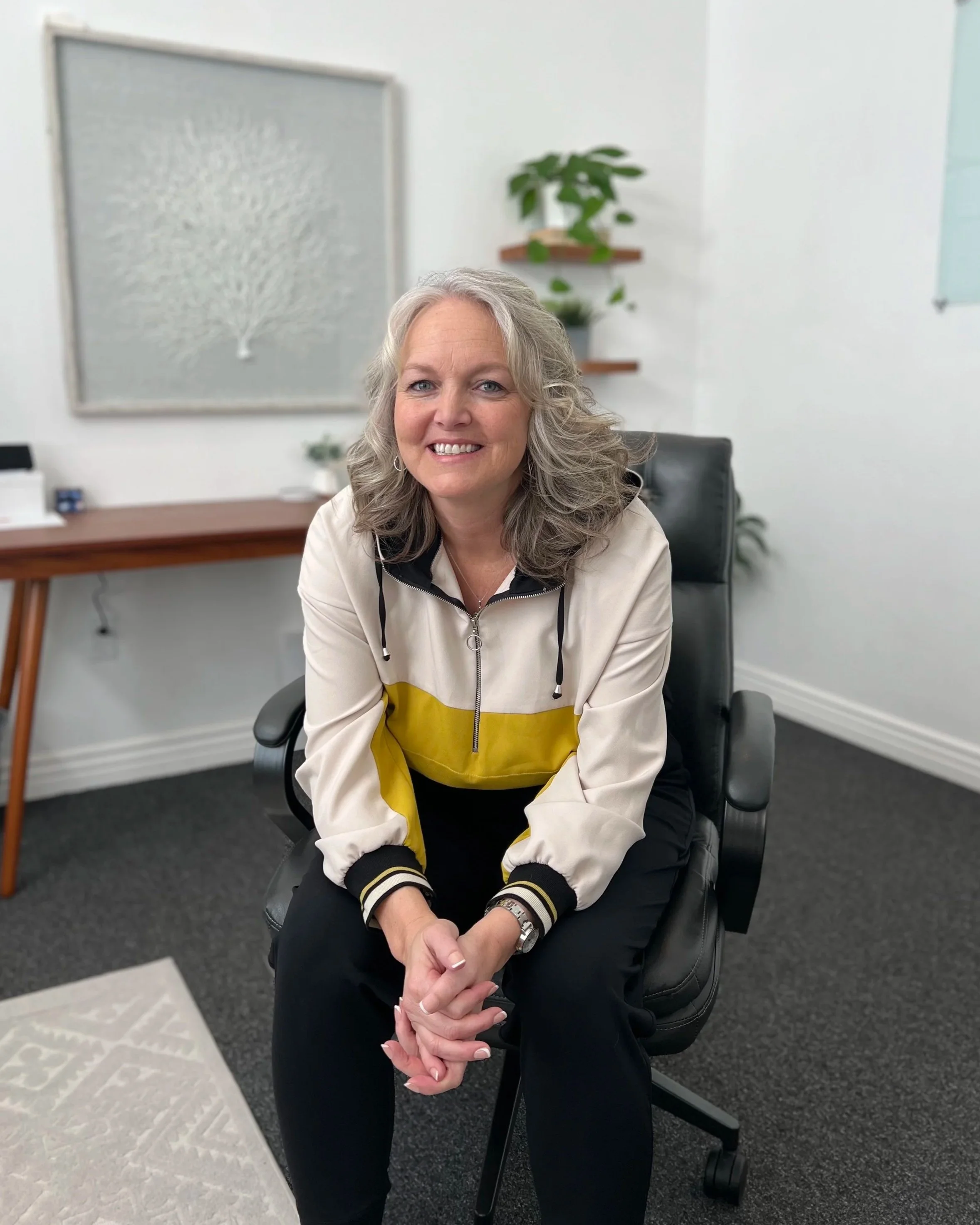A smiling woman wearing a beige and yellow jacket, sitting in an office chair in a modern office space with white walls, a black carpet, a office desk, and a wall art piece in the background.