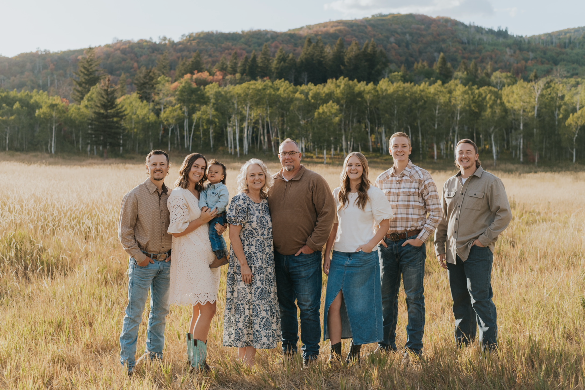 A family of eight standing in a grassy field with a forest and mountains in the background.