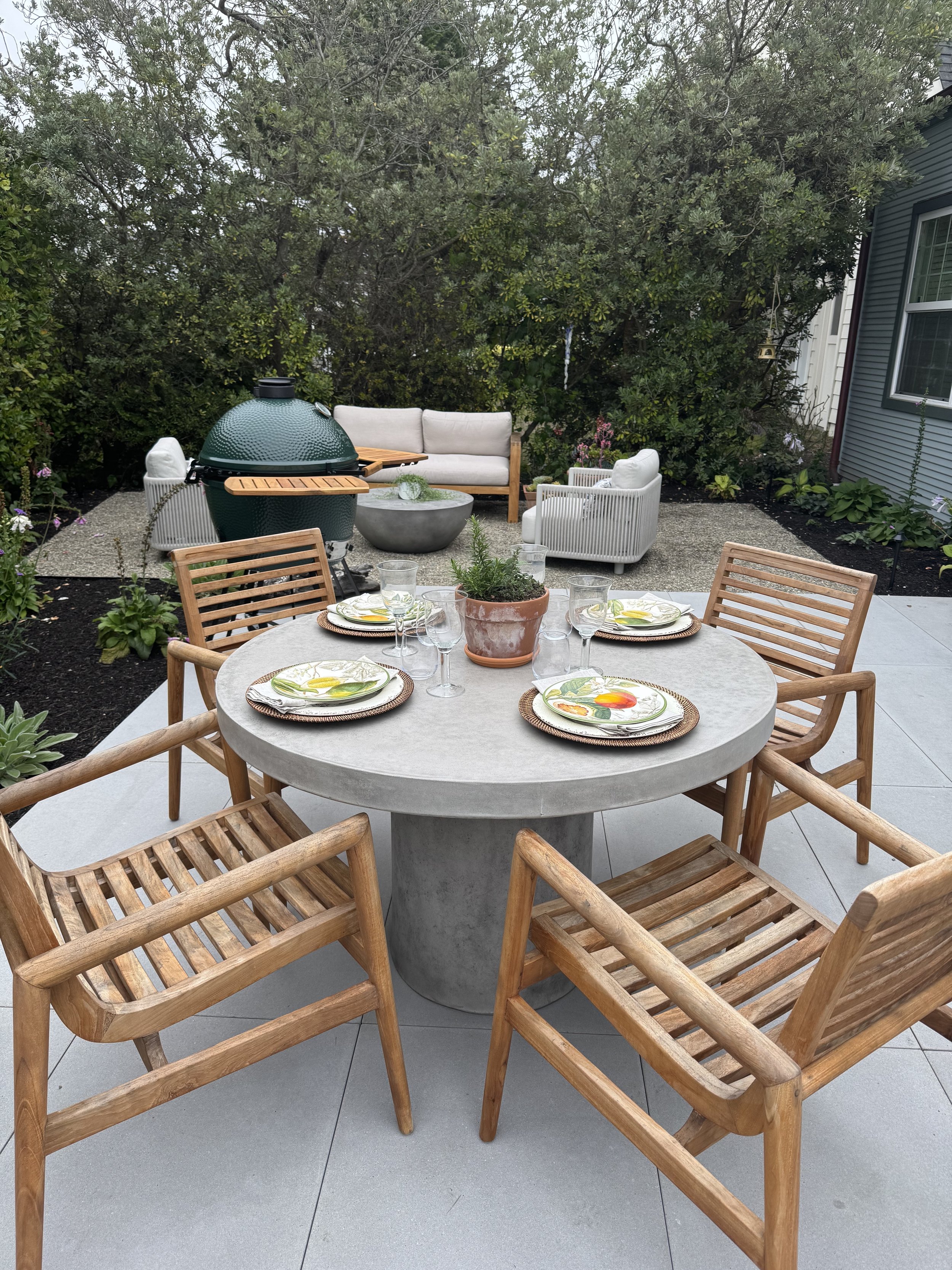 An outdoor patio with a round table set with plates and glasses, surrounded by wooden chairs, and a seating area with a couch and chairs in front of dense greenery.