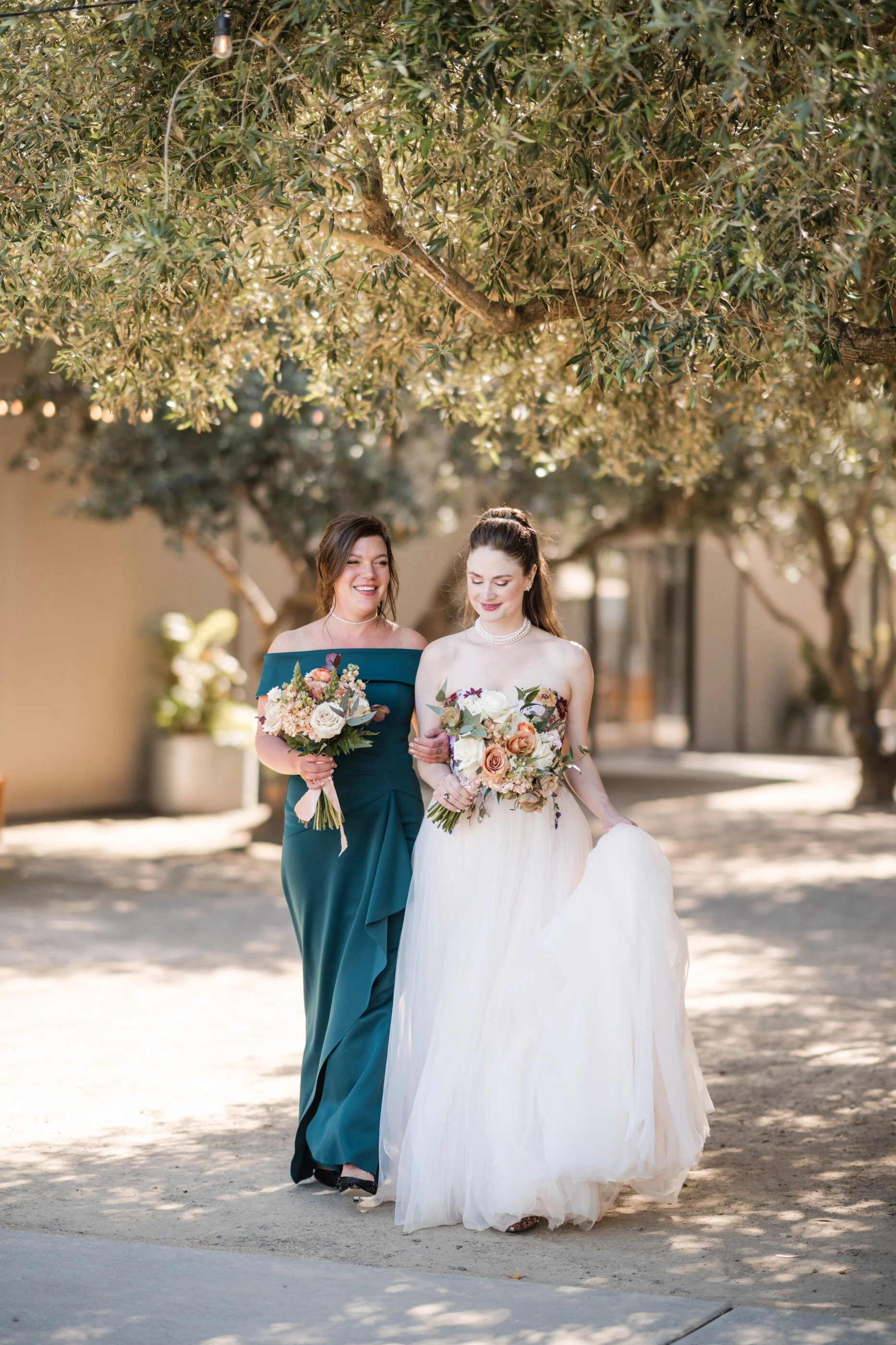 Fried walking with her bridesmaid during a romantic garden wedding, the bride wear soft glam bridal makeup with peach tones, glowing skin and half up bridal hairstyle with soft waves paired with a floral embroidered wedding gown and bouquet made of h