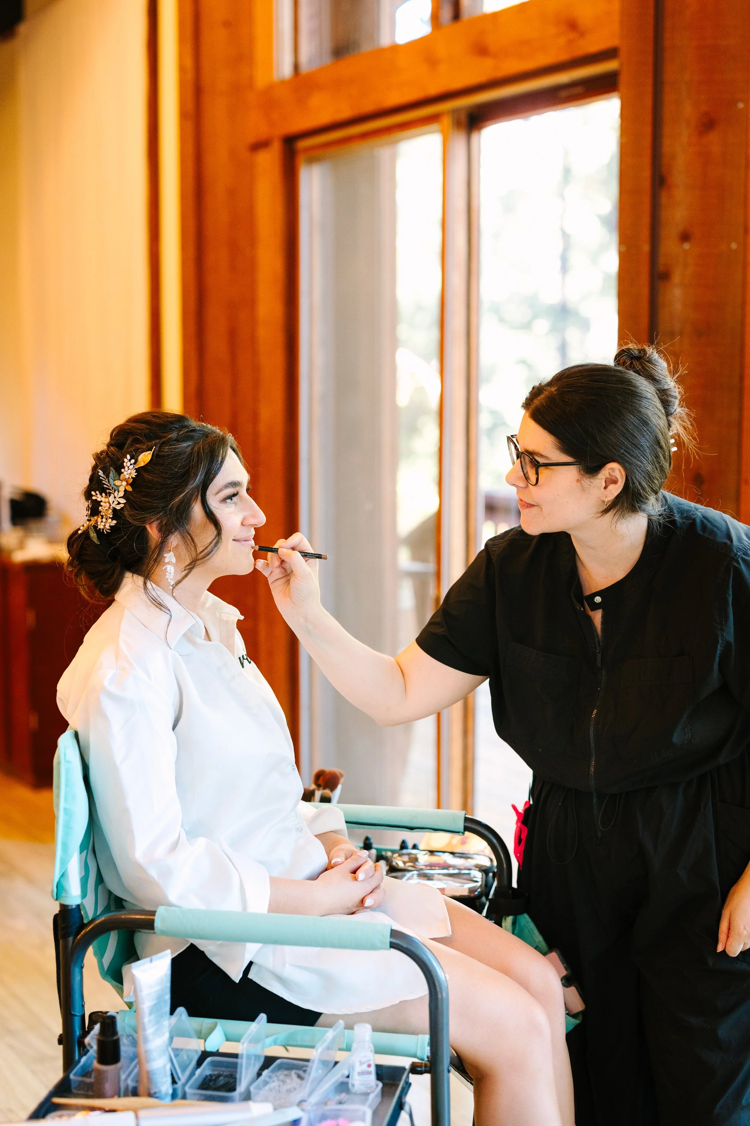 Make up artist applying natural bridal lip color during a redwood forest, wedding featuring soft glam, bridal makeup and romantic bridal updo by Beauty by Santi. Northern California soft textured up style bridal crown natural makeup behind the scenes