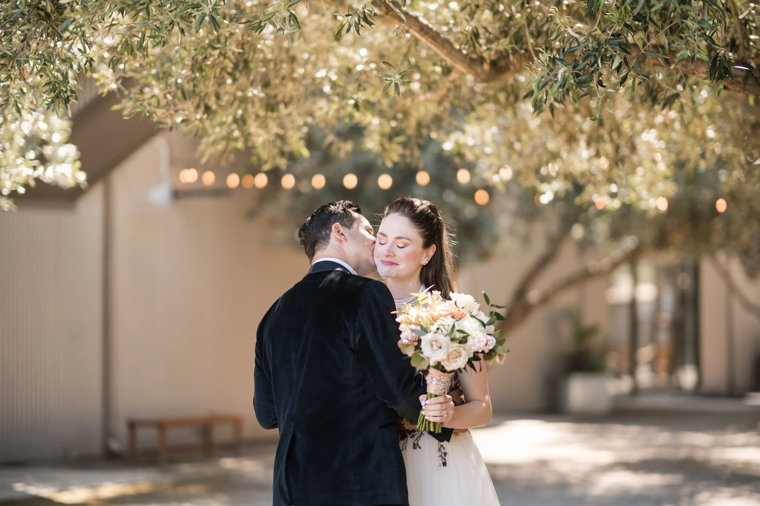 Romantic garden wedding moment with groom, kissing brides, cheek, bride feature, soft, glam, bridal makeup with peach tones, and half a bridal hairstyle with soft waves holding a romantic peach and bouquet under olive trees. Bridal color analysis war