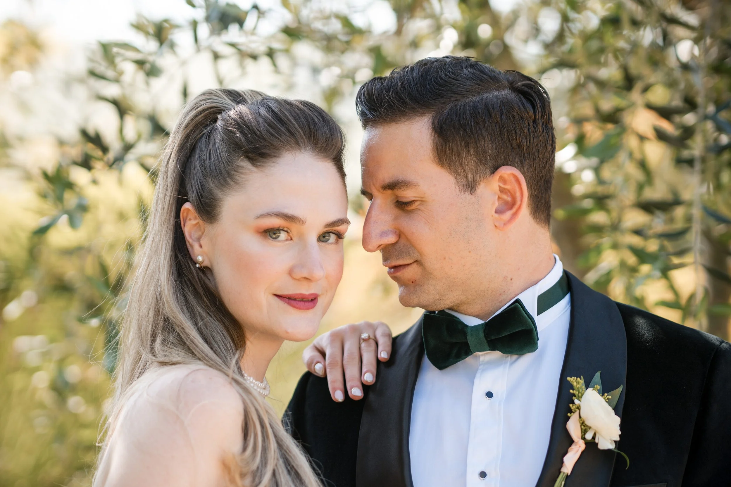 Romantic close-up portrait of bride and groom and garden, setting with groom in black tuxedo and bride, wearing pearl necklace and floral embroidered wedding gown. Spring bridal makeup, natural bridal makeup, soft, autumn bridal makeup. Bride wearing