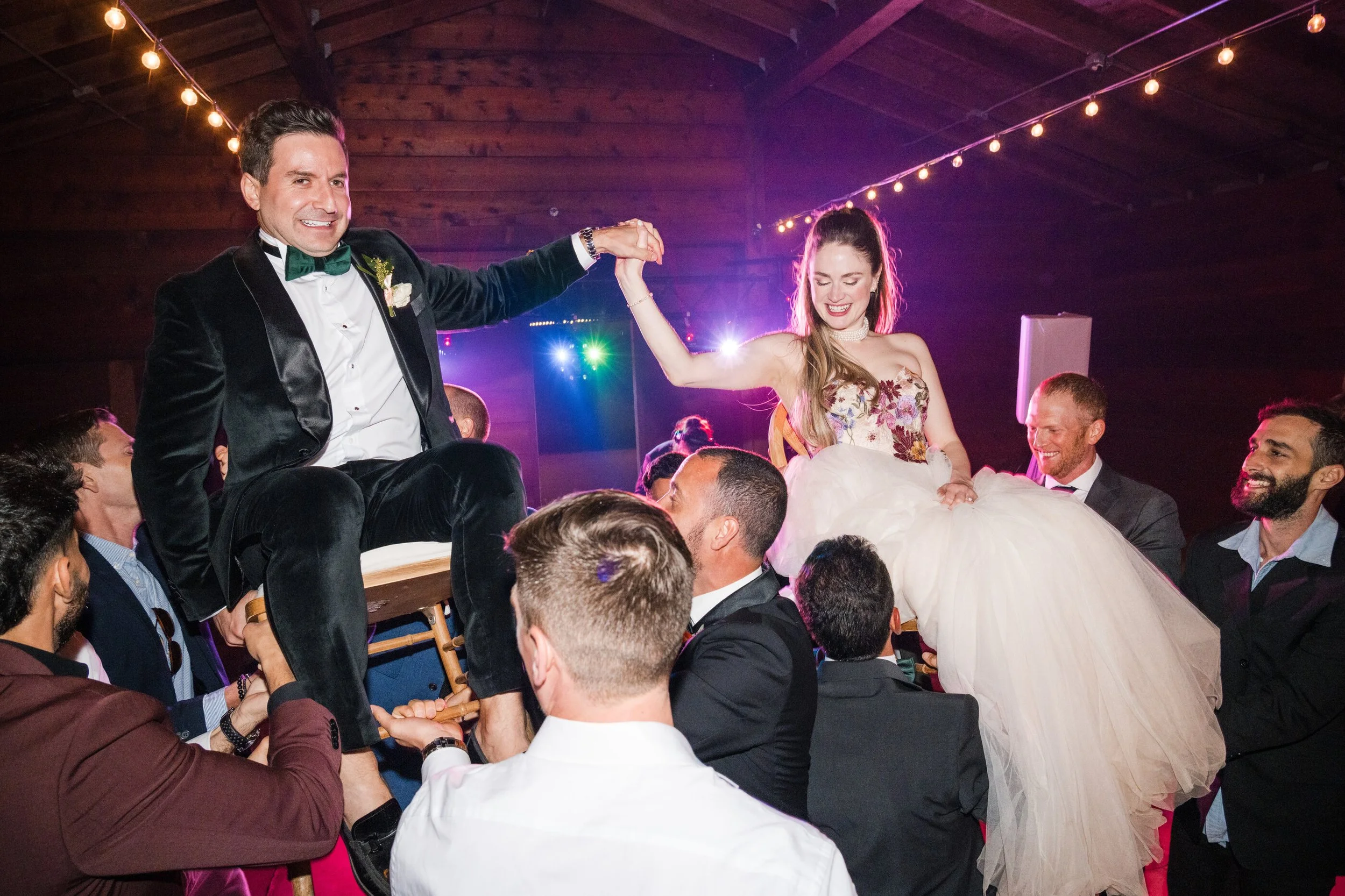 Bride and groom celebrating during their wedding reception as guest lift them on chairs while dancing under string lights, the bride wear soft glam bridal make up with glowing skin, warm, peach tones, and a romantic half of bridal hairstyle with soft