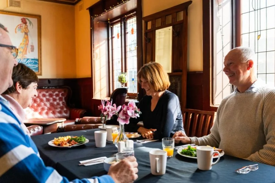 Group of five people having breakfast or brunch at a restaurant table, with flowers and sunlight streaming through stained glass windows.