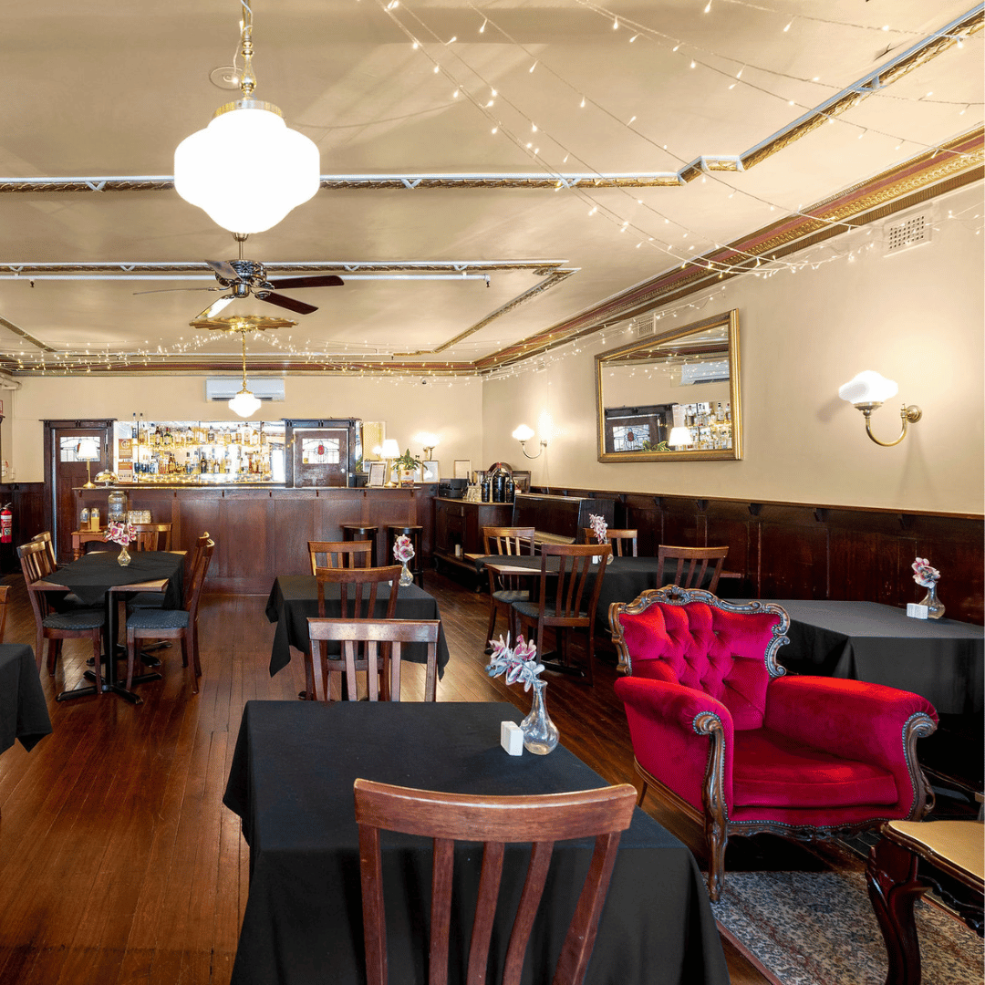 Elegant restaurant interior with black tablecloth-covered tables, wooden chairs, a red Victorian-style armchair, flowers in vases, and string lights on the ceiling.
