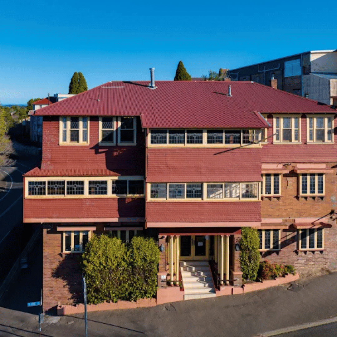 An aerial view of a multi-story brick building with a red-tiled roof and beige window frames, with trees and a clear blue sky in the background.