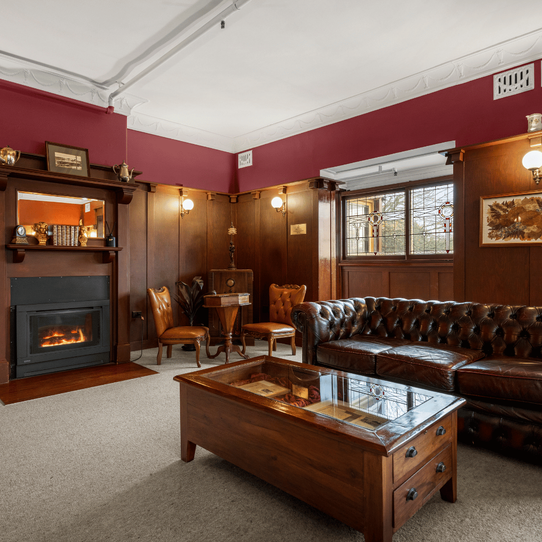 A cozy vintage living room with dark wood paneling, a leather sofa, a wooden coffee table with a glass top, a fireplace, and small decorative items on shelves.