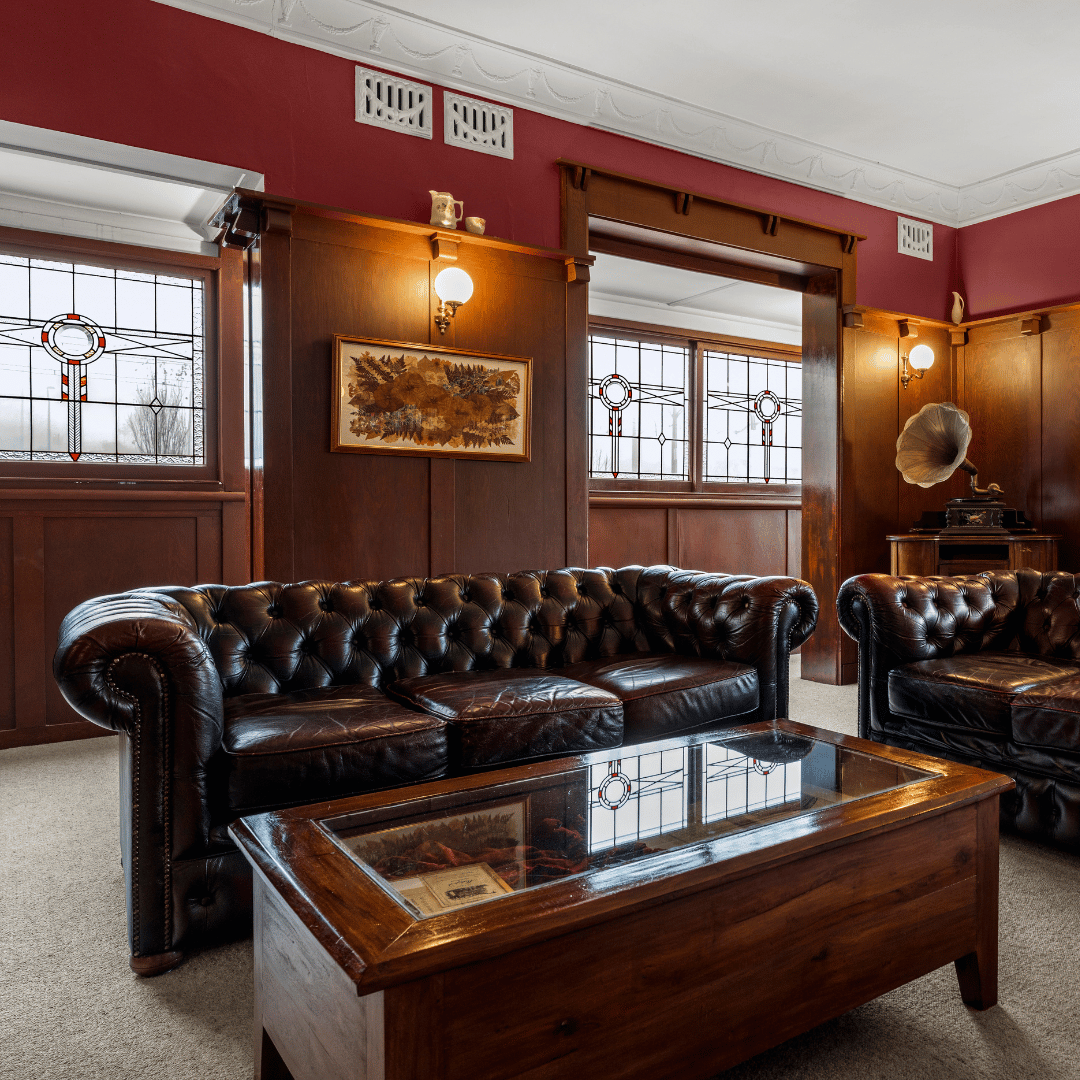 A vintage living room with dark wood-paneled walls, stained glass windows, leather Chesterfield sofas, a wooden coffee table with a glass top, and a vintage gramophone, illuminated by wall-mounted lamps.