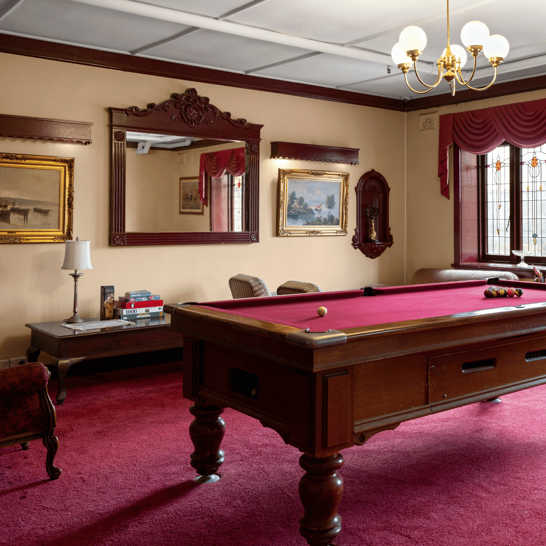 A vintage-style game room with a red pool table, framed paintings, a mirror, a table with books, chairs, a window with stained glass, and a chandelier ceiling light.