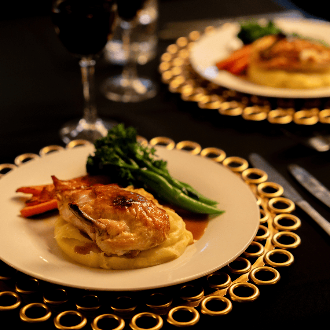 A plated serving of roasted chicken with mashed potatoes, broccoli, and carrots on a white plate with a gold decorative rim, set on a black table with a glass of red wine in the background.
