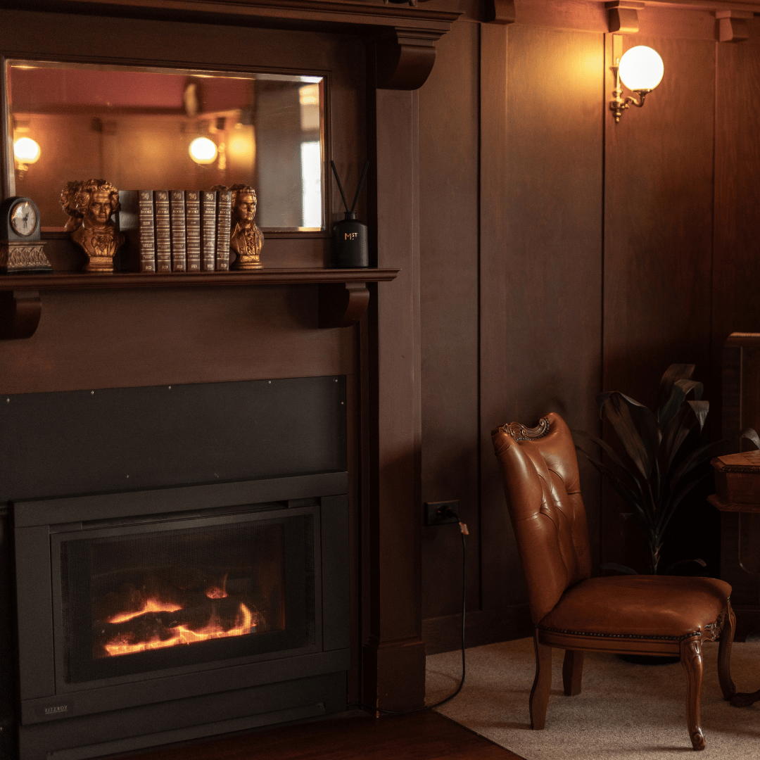 A cozy room with dark wooden paneling, a fireplace with a fire inside, a leather chair, a plant, a mirror above the fireplace, and decorative books and statues on the mantel