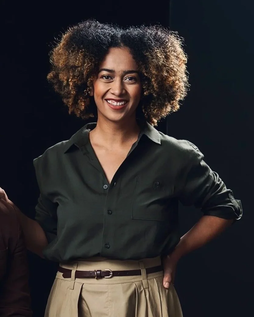 A woman with curly blonde hair wearing a red, long-sleeved blouse with a small white pattern, smiling, against a dark background.