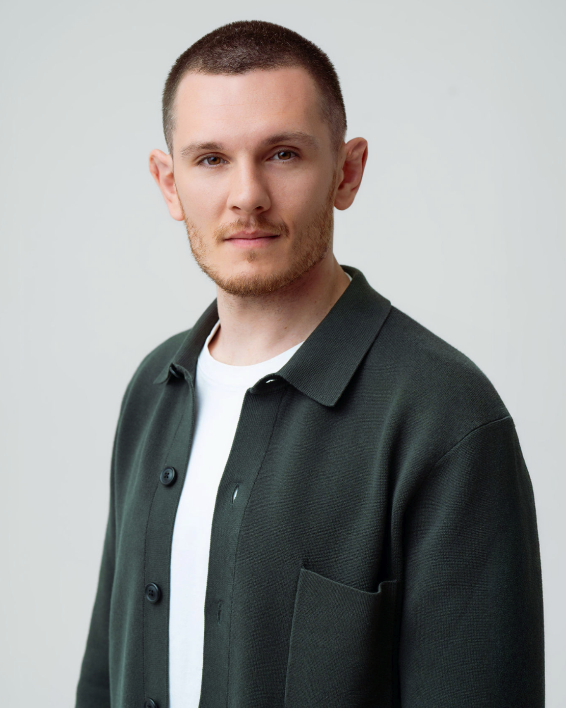 A young man with brown hair and a light mustache wearing a cream-colored sweater with orange and gray stripes on the chest, looking directly at the camera with a neutral expression against a neutral background.