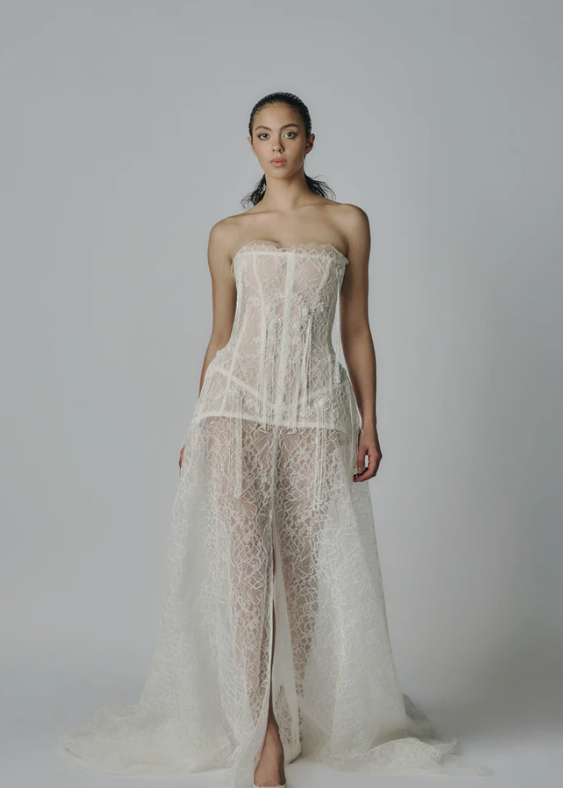 Australian Bride walking forwards with a white background and a sheer, fashionable, fashion forward and detailed lace gown with a structured corset.