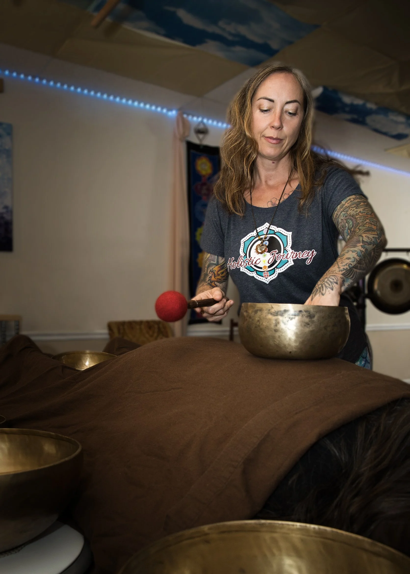 A woman with tattoos wearing a dark gray t-shirt, standing beside a massage table with a person lying face down, covered with a brown towel. She is holding a mallet and appears to be performing a sound therapy session in a decorated room with soft blue and white lighting.