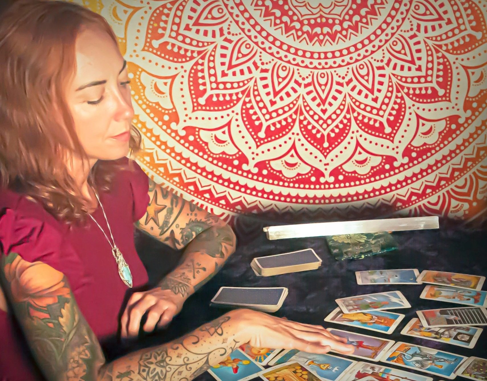 A woman with long, wavy, reddish-brown hair and colorful tattoos on her arms, sitting at a table with tarot cards spread out in front of her, with a large, intricate red and orange mandala on the wall behind her.