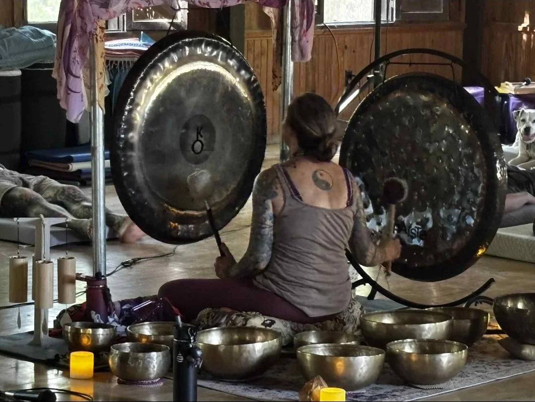 A woman with tattoos on her arms and a yin-yang tattoo on her back is sitting cross-legged, playing large singing bowls with mallets in a cozy wooden room. There are various smaller singing bowls, candles, and a tripod with additional instruments around her. A dog is lying down on a bed in the background, and another dog is sitting on a couch.