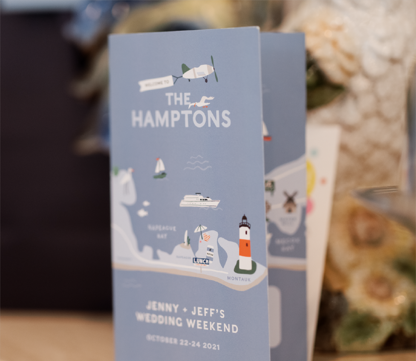 A blue wedding invitation card with illustrations of boats, a lighthouse, and a beach scene. Text reads "Welcome to The Hamptons," with details about Jenny and Jeff's wedding weekend from October 22-24, 2021.