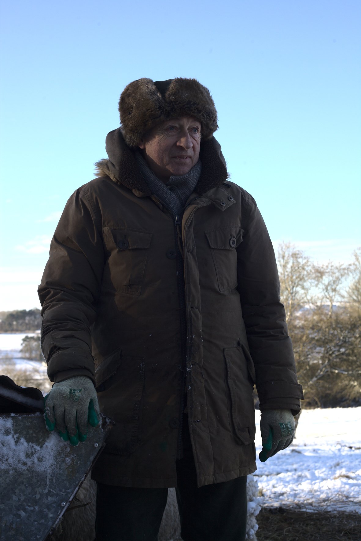 Portrait photograph by Hatty Frances Bell of a farmer wearing a big coat and hat standing in the snow in field with blue sky behind