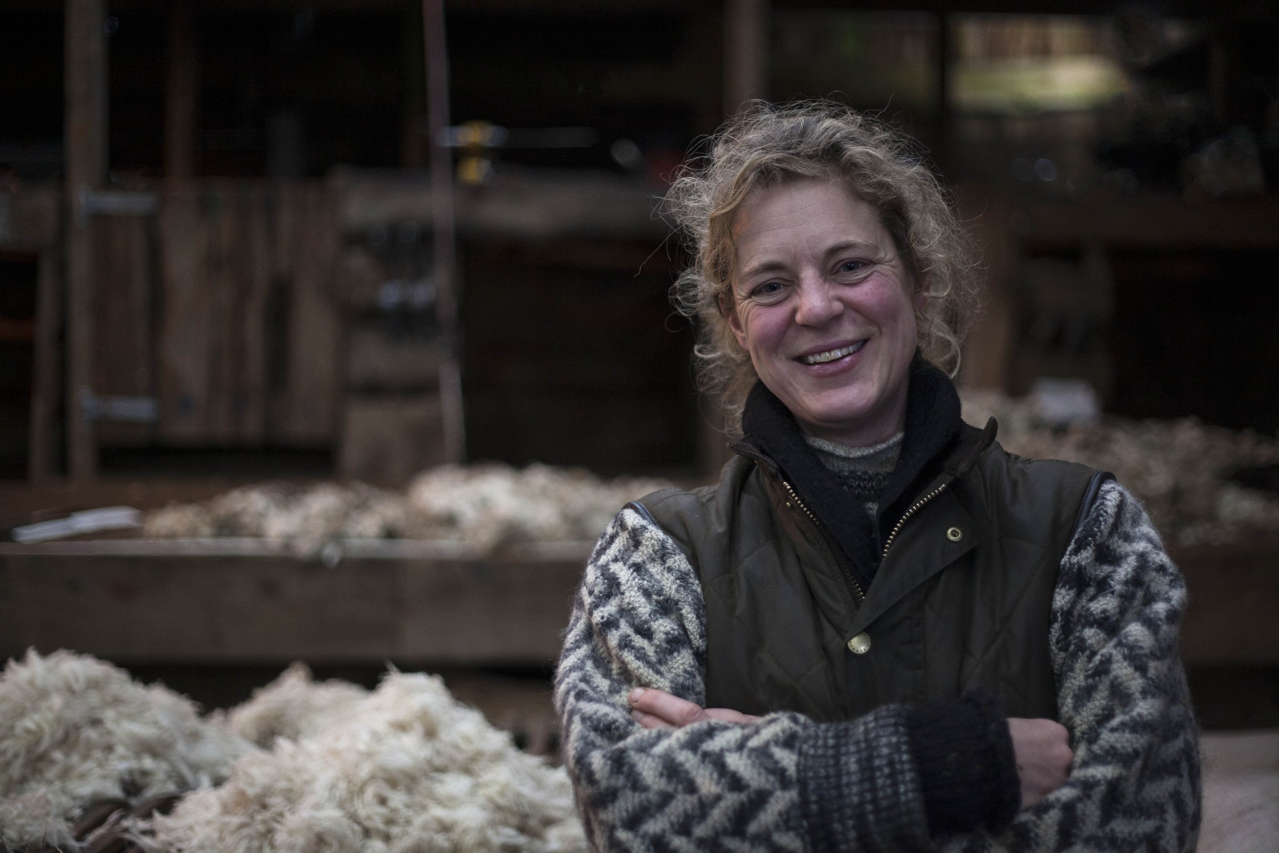Landscape photograph by Hatty Frances Bell of female farmer smiling in her barn full of wool at Fernhill Farm