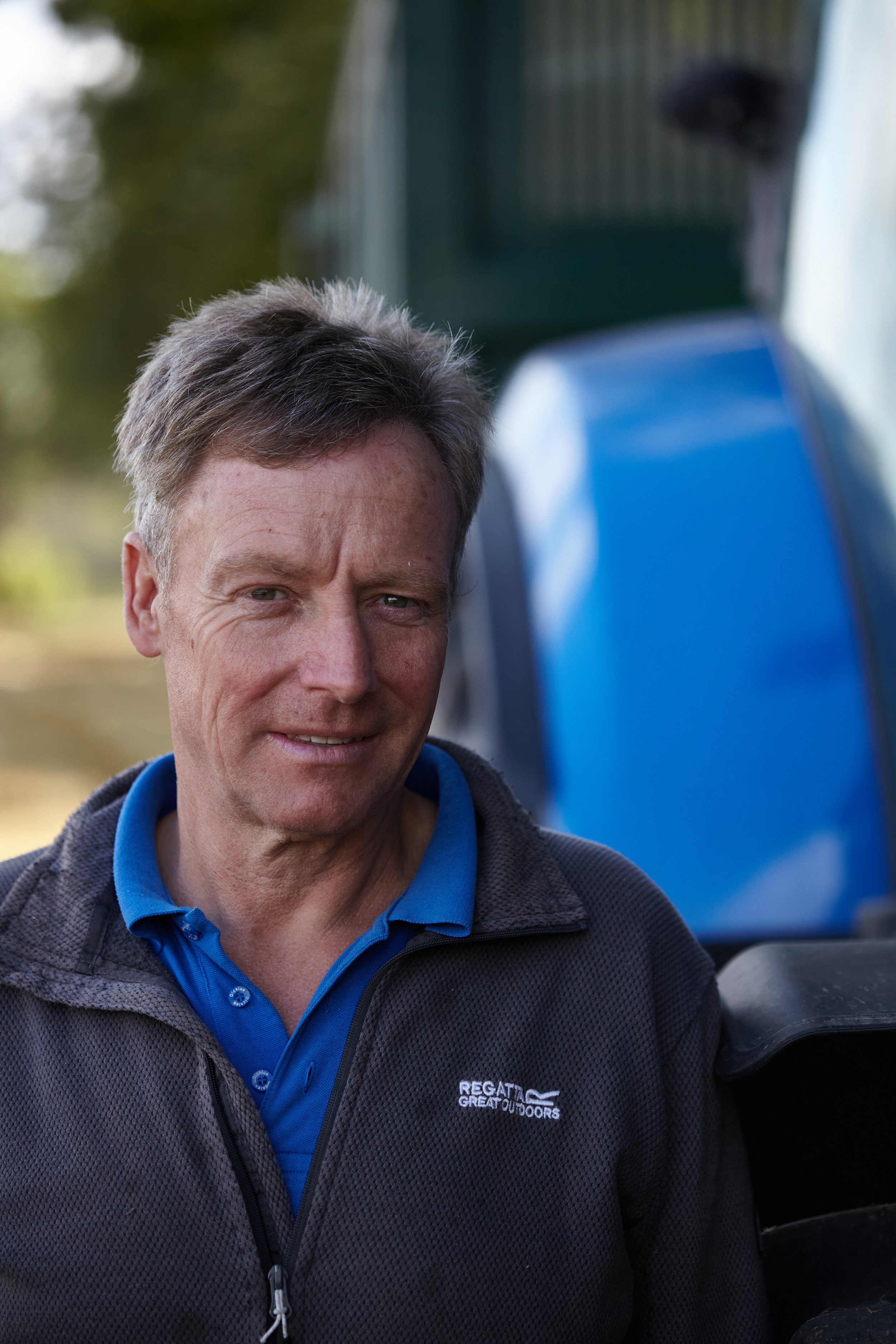 Headshot by Hatty Frances Bell taken in Norfolk of farmer standing next to his blue tractor in a field of cut wheat