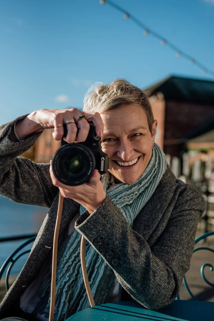 Dawn from DLT Visions is sitting at a table in the harbour. She is holding her camera up and smiling.
