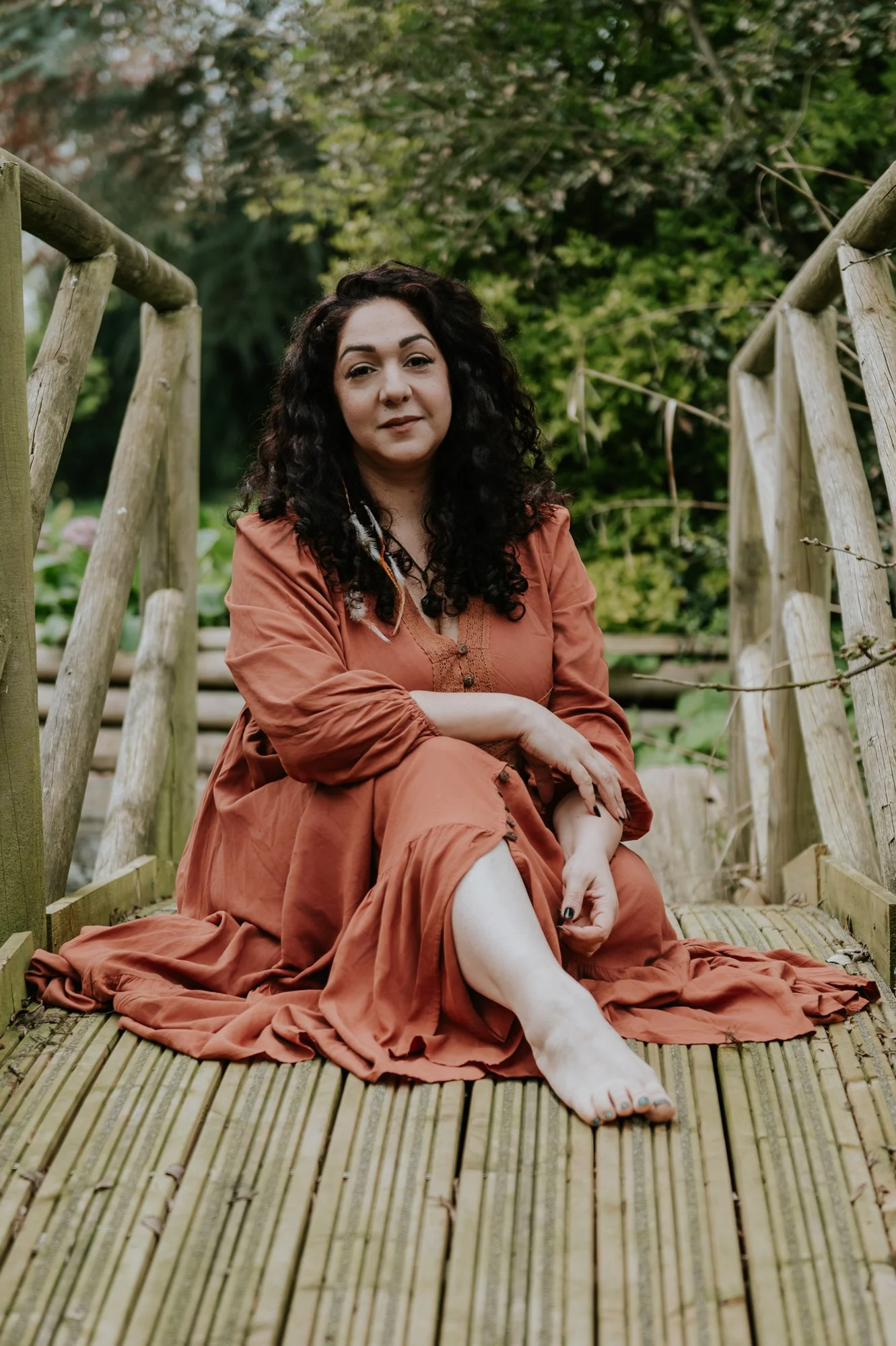 El Jedras, woman dressed in a flowy orange dress, sitting on a wooden bridge, she is looking relaxed and is gently smiling at the camera.
