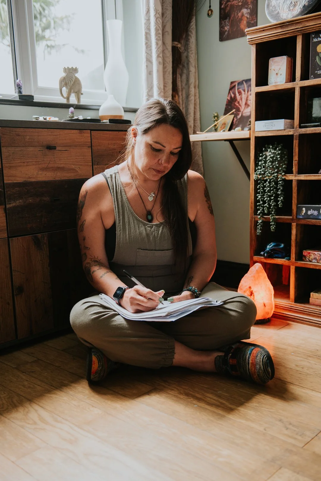 Alternative Advocacy. Abi is sitting cross legged on the floor. She is surrounded by beautiful wooden furniture filled with crystals and trinkets. She is writing in her notebook.