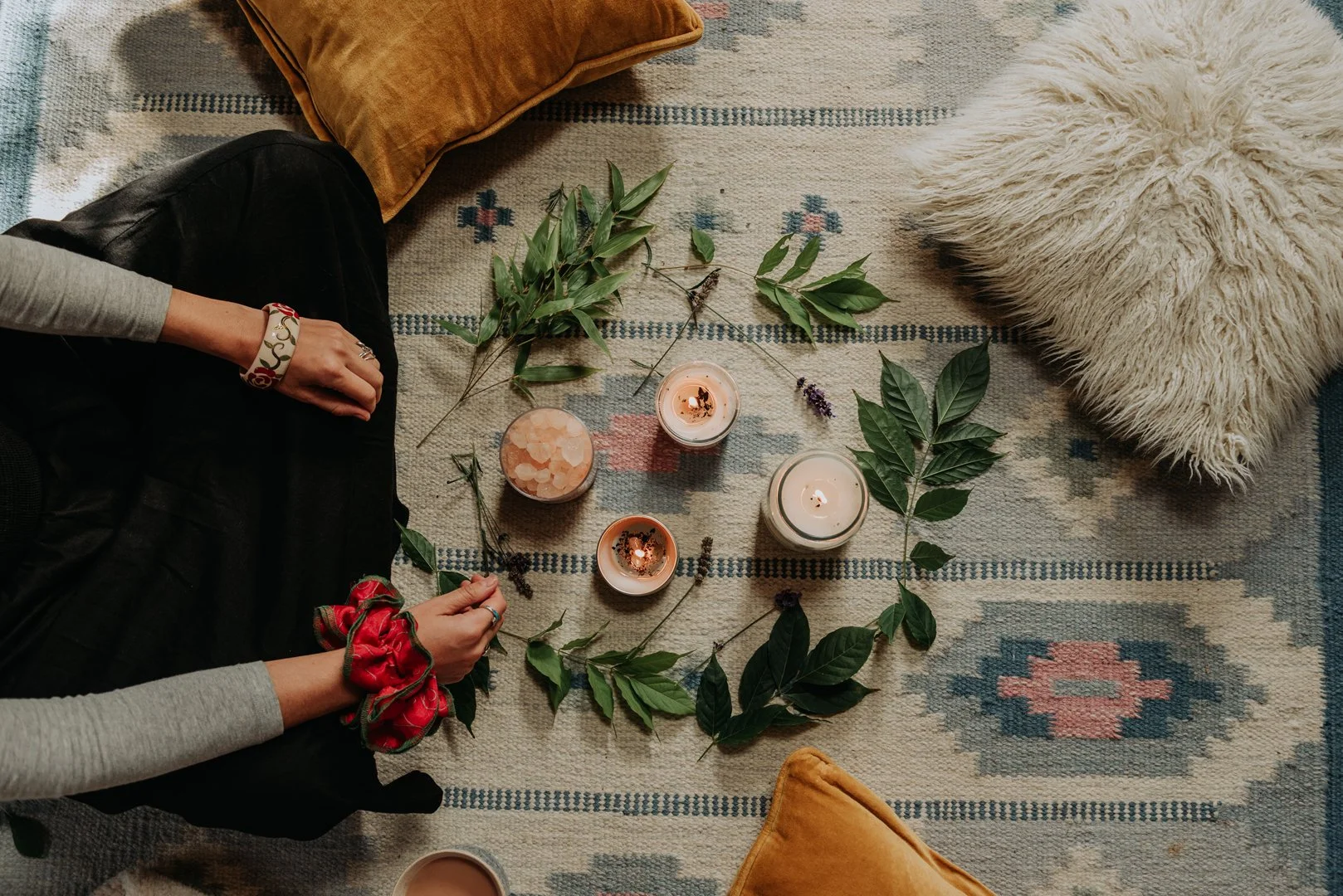 Looking down onto candles and crystals, there are pillows scattered around and arms are reaching into the left side ready for a women's circle.