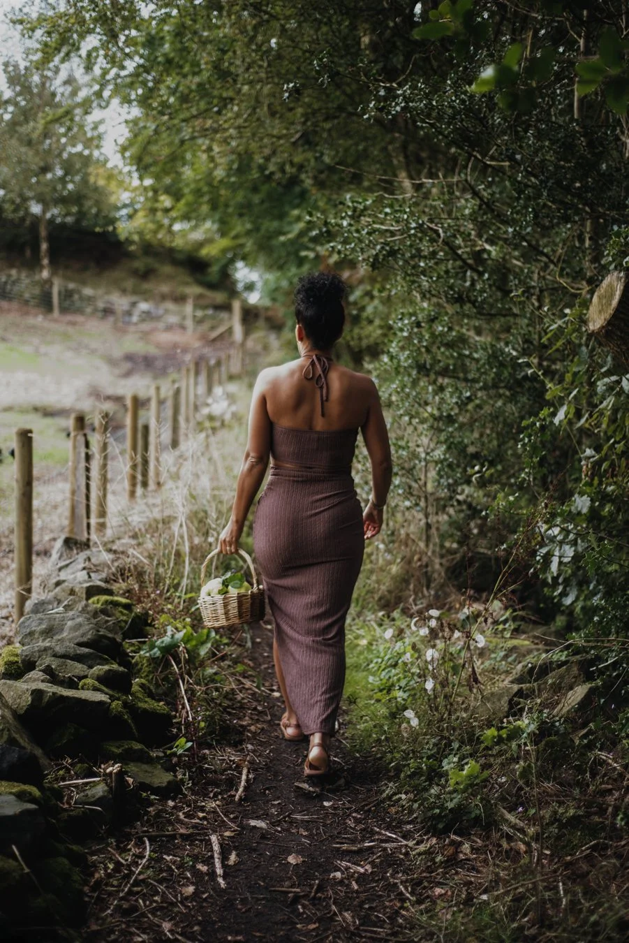 Woman walking away from camera through a woodland path. She is holding a basket to forage for plants