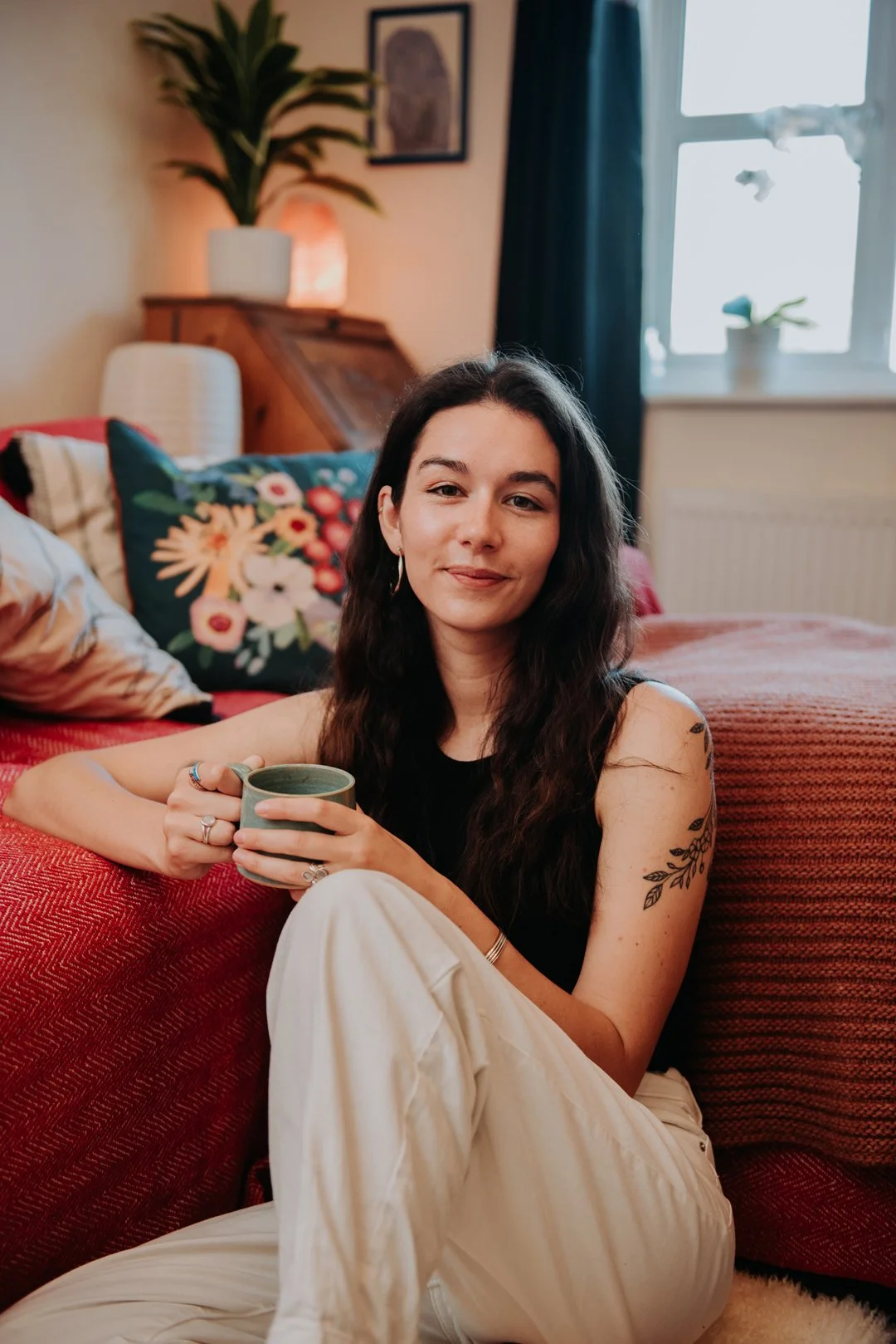 Ellie Belfield, therapist. She is sitting on the floor, leaning on her cosy sofa. She is holding a cup of herbal tea and smiling into the camera.