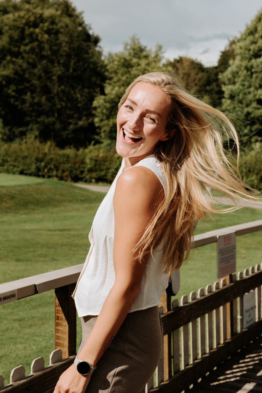 April Wild, Wild & Wild events. It is a bright summer looking day, April is turned away from the camera but is looking and smiling at the camera over her shoulder. Her long blonde hair is blowing in the wind behind her.