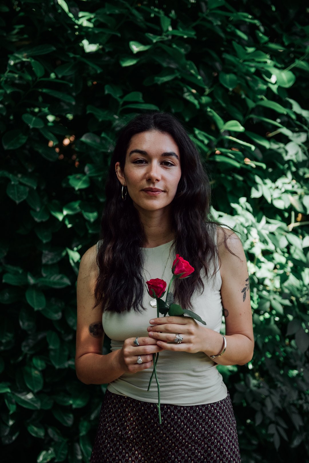 Ellie Belfield, Therapist. She is outdoors surrounded by lush greenery, she's holding two red roses and looking into the camera.